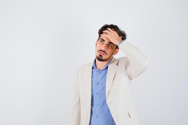 Jeune homme mettant la main sur le front, regardant loin en t-shirt bleu et veste de costume blanc et à l'air pensif