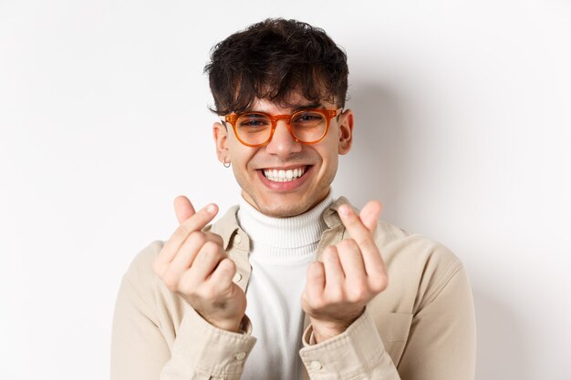 Jeune homme mignon dans des verres souriant et montrant des coeurs de doigt, debout sur fond blanc