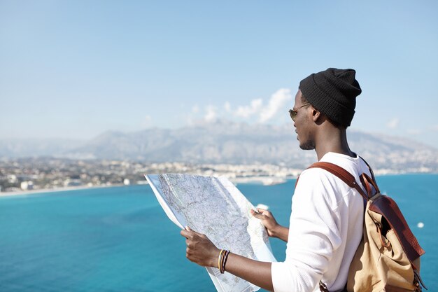 Jeune homme à la mode backpacker tenant une carte papier, debout au sommet de la montagne au-dessus de l'océan,