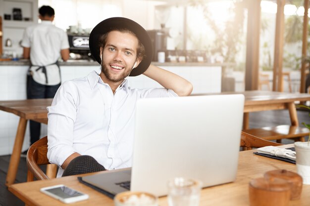 Jeune homme moderne en couvre-chef à la mode s'amuser seul, profiter du temps libre au café, naviguer sur Internet, utiliser le wifi gratuit sur ordinateur portable, écouter de la musique en ligne sur des écouteurs