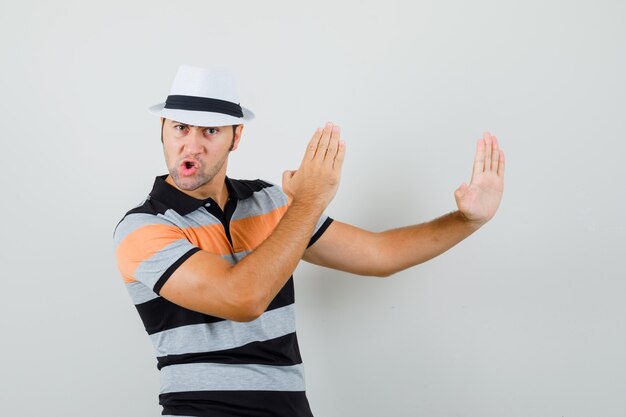 Jeune homme montrant la côtelette de karaté en t-shirt rayé, chapeau et à la vue flexible, avant.