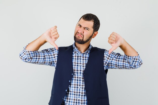 Jeune homme montrant deux pouces vers le bas en chemise, gilet et à la colère.