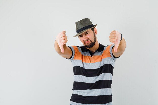 Jeune homme montrant deux pouces vers le bas en t-shirt, chapeau et à l'insatisfaction