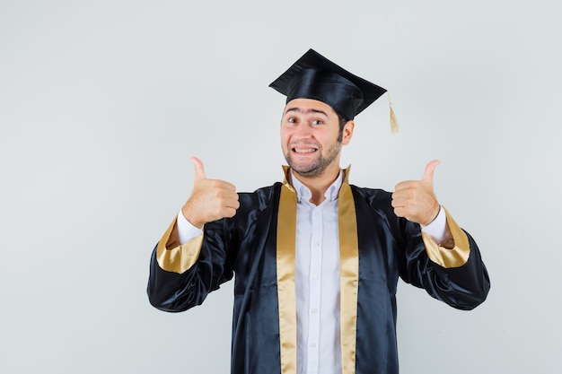 Jeune homme montrant deux pouces vers le haut en uniforme d'études supérieures et à la joyeuse. vue de face.