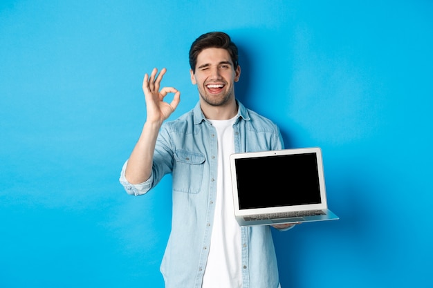 Jeune homme montrant un écran d'ordinateur portable et signe d'accord, approuver ou aimer la promo sur Internet, souriant satisfait, debout sur fond bleu