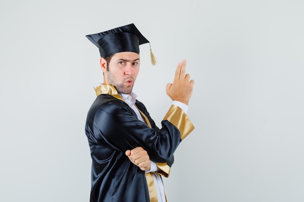 Jeune homme montrant le geste des armes à feu en uniforme diplômé et à la confiance. .