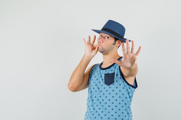 Jeune homme montrant le geste d'arrêt avec la bouche fermée comme zip en maillot bleu, chapeau et à la peur, vue de face.