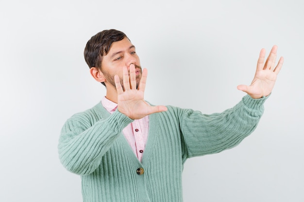 Jeune homme montrant un geste d'arrêt en chemise, cardigan et semblant résolu, vue de face.