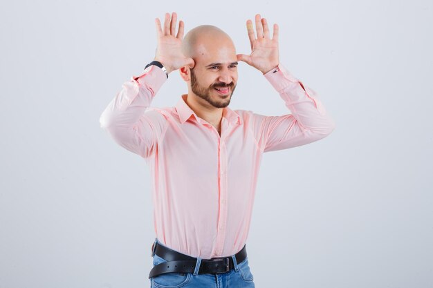 Jeune homme montrant un geste de corne de taureau tout en tirant la langue dans une chemise rose, un jean et l'air drôle, vue de face.