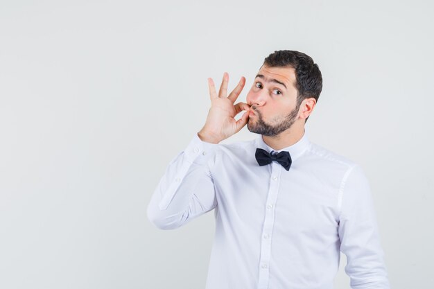 Jeune homme montrant un geste de fermeture éclair en chemise blanche et à la recherche de prudence. vue de face.