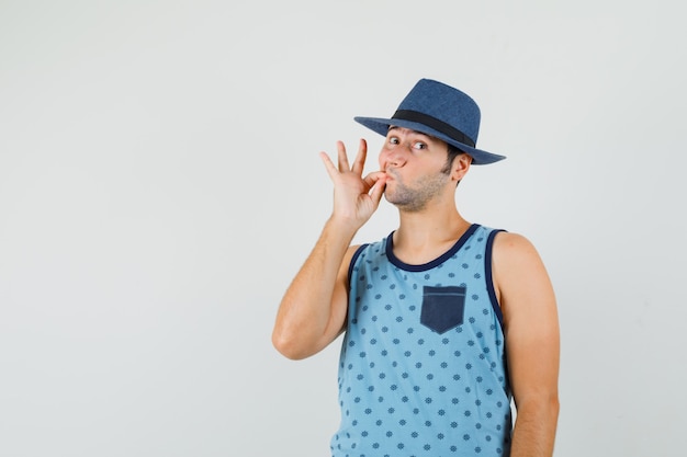 Photo gratuite jeune homme montrant le geste de fermeture éclair en maillot bleu, chapeau et à la vue attentive, de face.