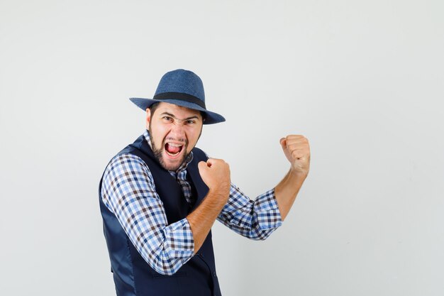 Jeune homme montrant le geste gagnant en chemise, gilet, chapeau et à la béatitude.