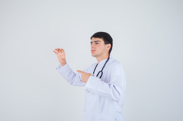 Jeune homme montrant le geste de la griffe, pointant vers le côté gauche en uniforme de médecin et à la joyeuse