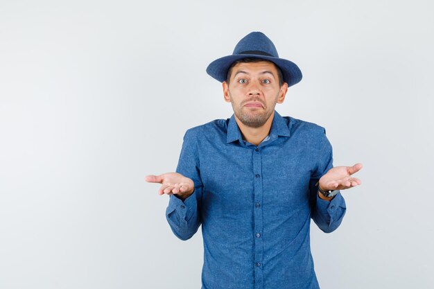 Jeune homme montrant un geste impuissant en chemise bleue, chapeau et l'air confus, vue de face.