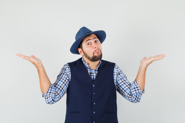 Jeune homme montrant un geste impuissant en chemise, gilet, chapeau et à la confusion.