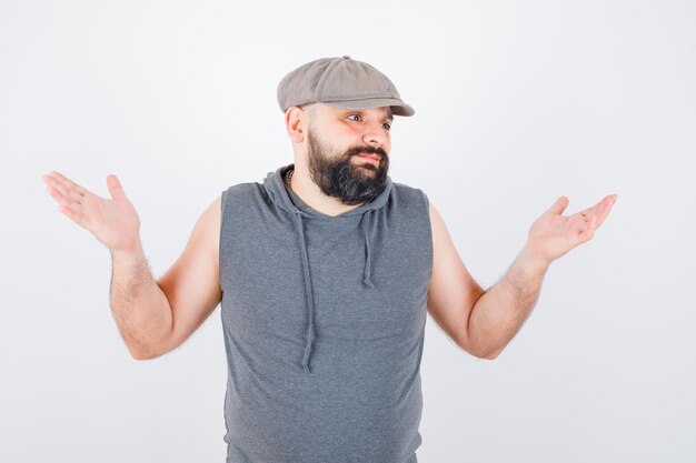 Jeune homme montrant un geste impuissant dans un sweat à capuche sans manches, une casquette et l'air désespéré. vue de face.