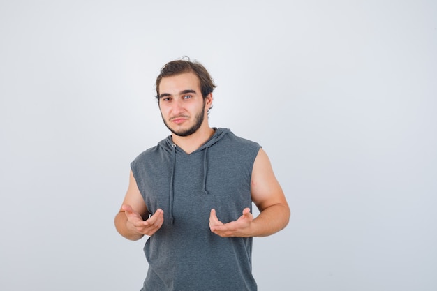 Jeune homme montrant un geste impuissant en sweat à capuche sans manches et à la perplexité. vue de face.