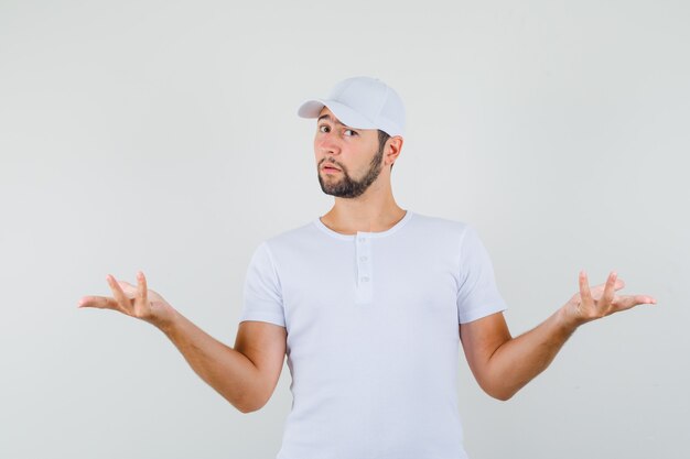 Jeune homme montrant un geste impuissant en t-shirt blanc, casquette et à la tristesse. vue de face.