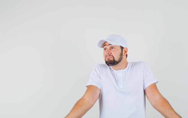 Jeune homme montrant un geste impuissant en t-shirt, casquette et à la vue troublée, de face. espace pour le texte