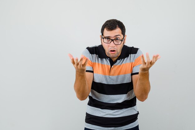 Jeune homme montrant un geste impuissant en t-shirt, lunettes et à la troublé. vue de face.