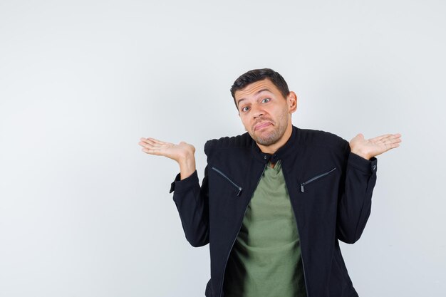 Jeune homme montrant un geste impuissant en t-shirt, veste et semblant mignon, vue de face.