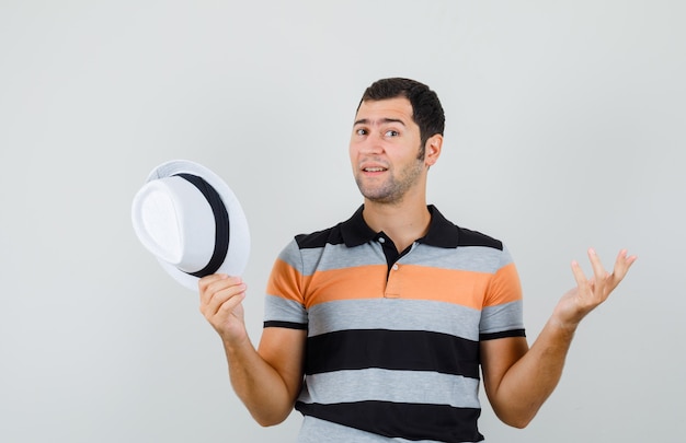 Jeune homme montrant un geste impuissant tout en tenant un chapeau en t-shirt, chapeau et à la perplexité. vue de face.