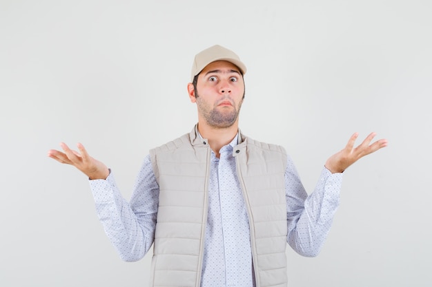 Jeune homme montrant un geste impuissant en veste beige et casquette et à la surprise, vue de face.