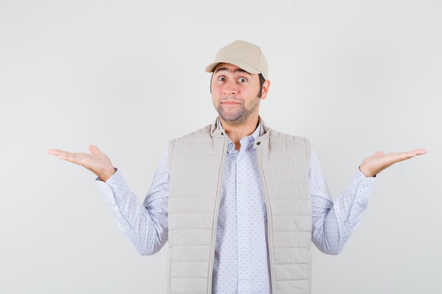 Jeune homme montrant un geste impuissant en veste beige et casquette et à la vue de face, heureux.