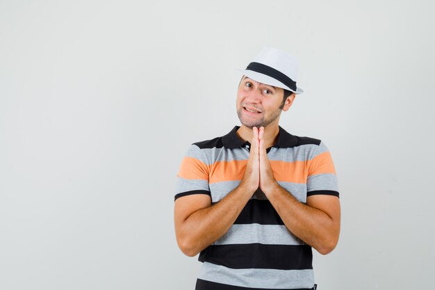 Jeune homme montrant le geste de namaste en t-shirt rayé, chapeau et à la vue positive, de face. espace pour le texte
