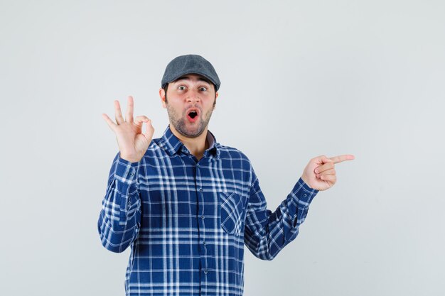 Jeune homme montrant un geste ok, pointant de côté en chemise, casquette et à la surprise. vue de face.