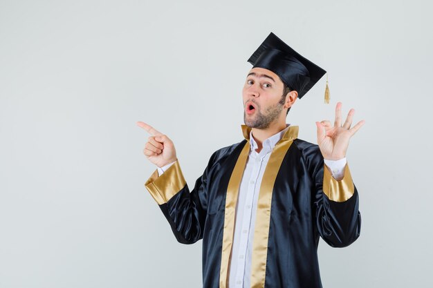 Jeune homme montrant un geste ok, pointant vers le haut en uniforme d'études supérieures et à la surprise. vue de face.