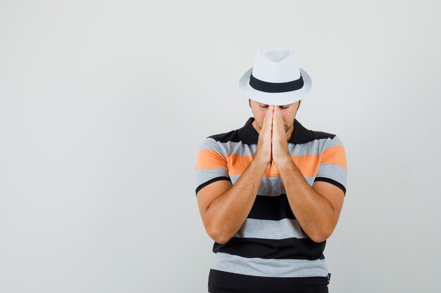 Jeune homme montrant le geste de prière en t-shirt rayé, chapeau et à la vue de face, concentré. espace pour le texte