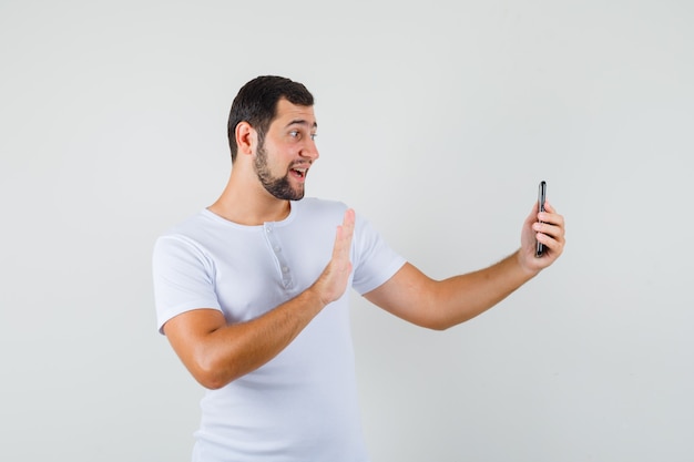 Jeune homme montrant le geste de salutation tout en faisant un appel vidéo en t-shirt blanc et à la recherche de plaisir.