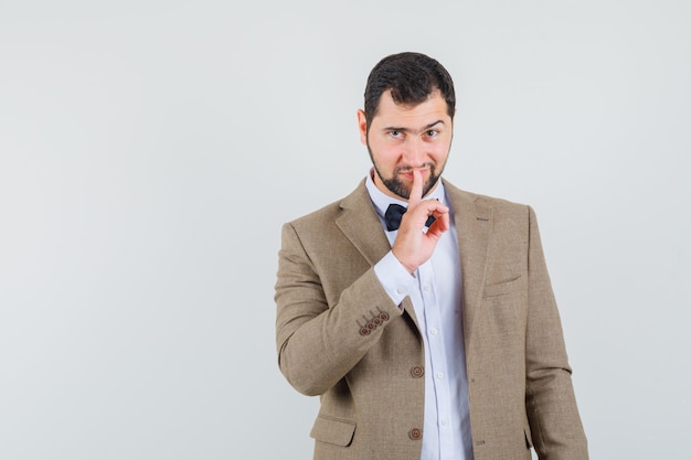 Jeune homme montrant le geste de silence en costume et à la ruse, vue de face.