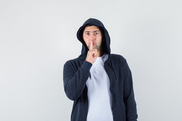 Jeune homme montrant le geste de silence en t-shirt, veste et à la recherche de sérieux. vue de face.