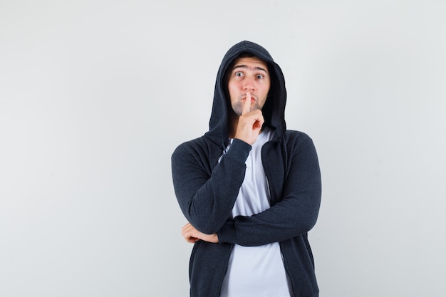 Jeune homme montrant le geste de silence en t-shirt, veste et regardant prudemment, vue de face.