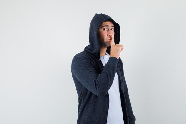 Jeune homme montrant le geste de silence en t-shirt, veste et regardant prudemment, vue de face.