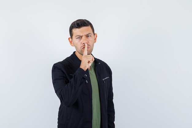 Jeune homme montrant un geste de silence en t-shirt, veste et semblant sérieux, vue de face.