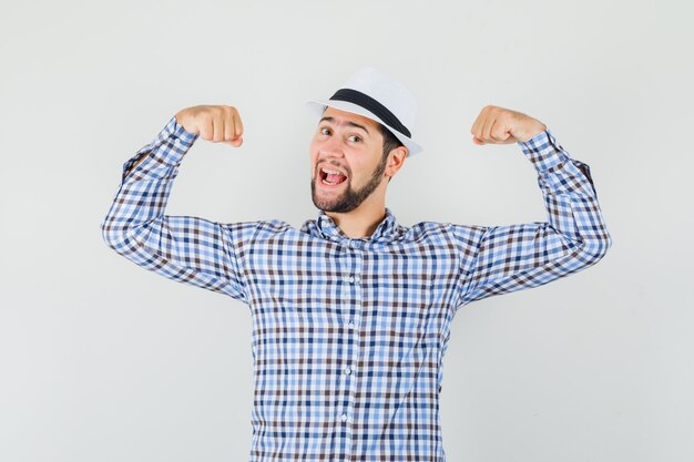 Jeune homme montrant les muscles des bras en chemise à carreaux, chapeau et à la fierté