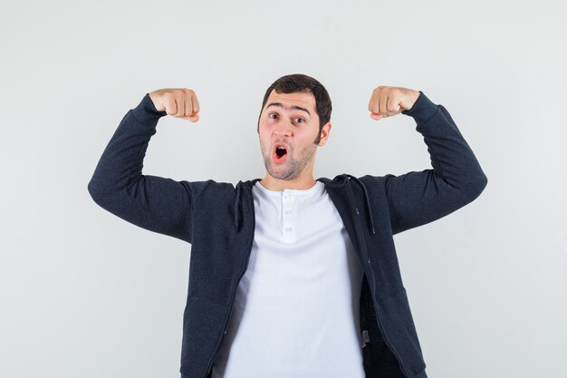 Jeune homme montrant des muscles en t-shirt blanc et sweat à capuche noir à glissière sur le devant et à l'optimiste, vue de face.