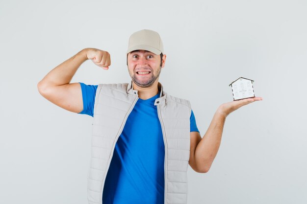 Jeune homme montrant les muscles, tenant le modèle de la maison en t-shirt, veste, casquette et à la chance, vue de face.