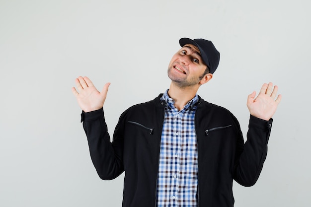 Jeune homme montrant des paumes dans un geste d'abandon en chemise, veste, casquette et à la joyeuse vue de face.