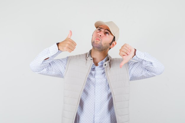 Jeune homme montrant le pouce de haut en bas en chemise, veste sans manches, casquette et à hésitant. vue de face.