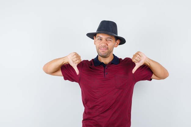 Jeune Homme Montrant Le Pouce Vers Le Bas En Chemise Rouge, Chapeau Noir Et L'air Mécontent, Vue De Face.