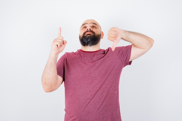 Jeune homme montrant le pouce vers le bas tout en pointant vers le haut en t-shirt rose, vue de face.