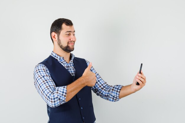 Jeune homme montrant le pouce vers le haut sur le chat vidéo en chemise, gilet et à la joyeuse.