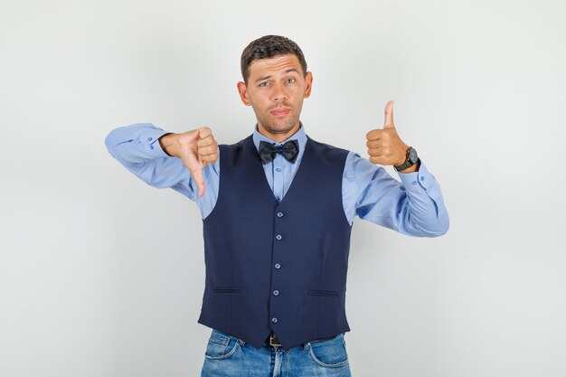 Jeune homme montrant les pouces de haut en bas en costume, jeans et à la confusion.