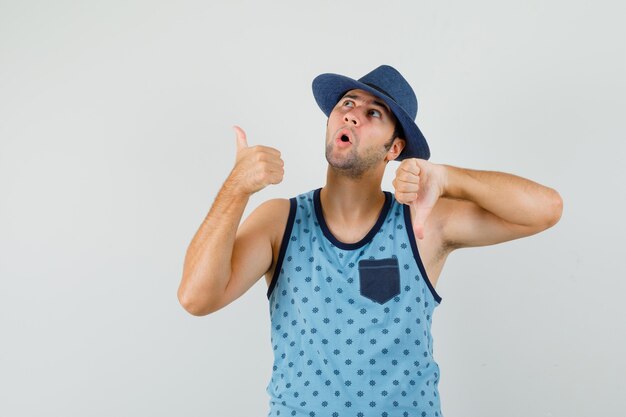 Jeune homme montrant les pouces de haut en bas en maillot bleu, chapeau et à la vue de face, hésitant.