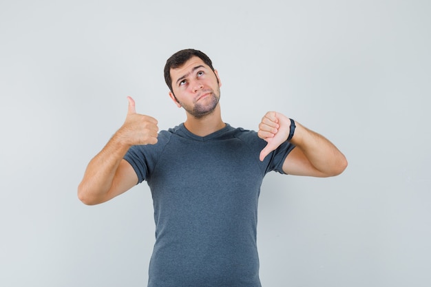 Jeune homme montrant les pouces de haut en bas en t-shirt gris et à la vue de face, hésitant.