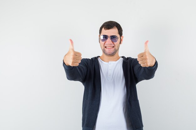 Jeune homme montrant les pouces vers le haut en t-shirt, veste et à la joyeuse, vue de face.